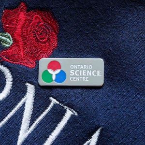 May include: A silver rectangular pin with the text "ONTARIO SCIENCE CENTRE" and a logo featuring red, green, and blue circles. The pin is set against a dark blue fabric with a red rose embroidery and white stitching.