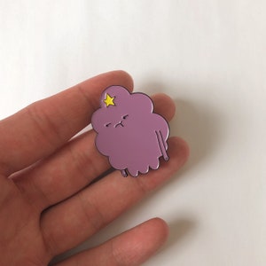 May include: A purple and yellow enamel pin of a cartoon character with a grumpy expression. The character has a star on its head and is shaped like a cloud.