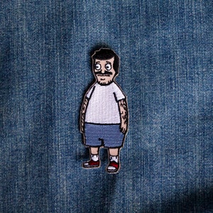 Peut inclure: Patch brodé représentant un personnage de dessin animé avec des cheveux noirs, une chemise blanche, un short bleu et des chaussures rouges. Le personnage a une moustache et une expression surprise.