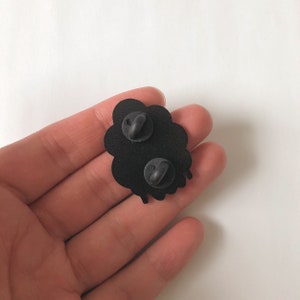 May include: Black enamel pin shaped like a cloud with dripping edges. The pin has two metal clasps on the back.