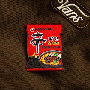 May include: Red and black embroidered patch of a Shin Ramyun instant noodle packet. The packet has the brand name "Nongshim" and the Korean text "라면 Shin Noodle Soup".