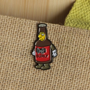 May include: An enamel pin of a Duff beer bottle with a cartoon face and the text "Duff Surly".