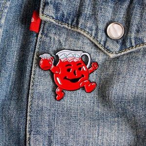 Puede incluir: Pin de esmalte rojo de un lanzador sonriente que sostiene un vaso de líquido rojo con cubitos de hielo. El lanzador tiene una tapa blanca y una cara blanca con una sonrisa y ojos rojos. El pin está en una chaqueta vaquera azul.