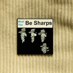 May include: Black and white enamel pin featuring a cartoon band called "The Be Sharps". The pin has a white background with black text that reads "Meet The Be Sharps".