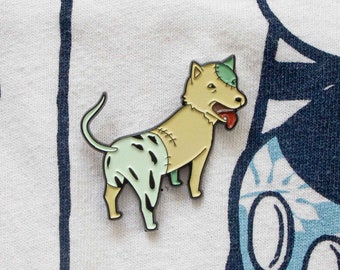 May include: A green, white, and brown enamel pin of a dog with a stitched-up face. The dog is standing on all fours with its tongue sticking out.