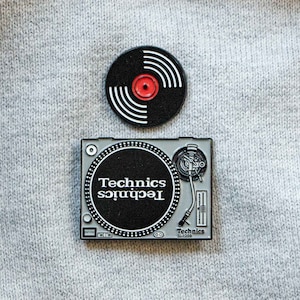 May include: A silver and black enamel pin of a turntable with the text "Technics" and "Technics SL-1200" on it. A separate black and white enamel pin of a vinyl record is also included.