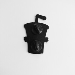 May include: Black enamel pin of a cartoon cup with a straw. The pin has two black metal clasps on the back.