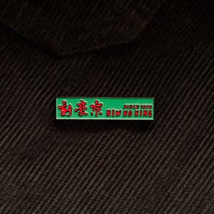 May include: Green and red enamel pin with the text "New Ho King" and Chinese characters. The pin also says "Since 1976".