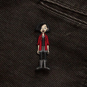 May include: Embroidered patch of a cartoon woman with black hair, wearing a red jacket, black trousers, and black boots.