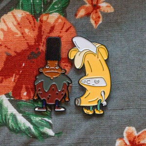 May include: Two enamel pins featuring cartoon characters. The first pin is of a brown-skinned character with a green shirt and brown pants. The second pin is of a yellow banana with a white face and arms.