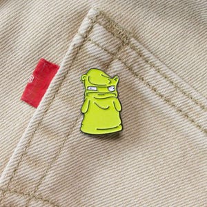 May include: A yellow enamel pin featuring a cartoon character with a green face and a yellow body. The character has a surprised expression with its eyes wide open and its mouth closed. The pin is displayed on a light brown fabric background.