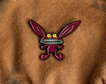 May include: A red cartoon character with large ears, yellow eyes, and a wide, toothy grin. The character is standing on a brown fabric background.