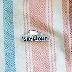 May include: A white enamel pin with a blue outline depicting the SkyDome, a domed stadium in Toronto, Canada. The pin features the word "SKYDOME" in blue letters.
