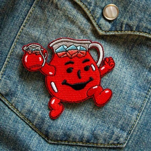 May include: Red embroidered patch of a smiling cartoon pitcher holding a smaller pitcher. The pitcher is wearing a red, white and blue hat and has a red, white and blue drink inside. The patch is on a blue denim background.