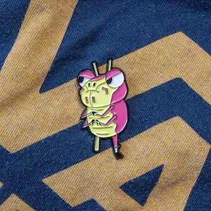 May include: A pink and yellow cartoon bug enamel pin with angry eyes and crossed arms. The pin is on a blue and yellow fabric background.