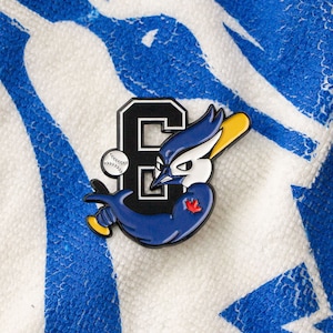 Peut inclure: Une épingle en émail bleu et noir représentant un geai bleu de dessin animé tenant une batte de baseball avec un ballon de baseball en arrière-plan. Le geai bleu porte un érable rouge sur la poitrine et le numéro "6" est derrière lui.