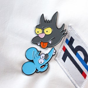 May include: Two enamel pins featuring cartoon characters. The first pin is a grey cat with yellow eyes and a pink nose. The second pin is a blue mouse with large eyes and a pink nose.