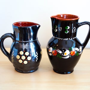 May include: Two black ceramic pitchers with painted designs. One pitcher has a white and red floral design, the other has a white and orange geometric design.