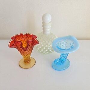 Könnte beinhalten: Eine Sammlung aus drei Glasobjekten: eine orange-gelbe Hobnail-Vase mit gewelltem Rand, eine klare weiße Hobnail-Karaffe mit rundem Stopfen und eine hellblaue Hobnail-Vase in Stiefelform mit einer breiten, ausgestellten Öffnung.