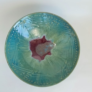May include: A large, round ceramic bowl with a teal glaze and a red and green center. The bowl has a textured surface with a subtle pattern.