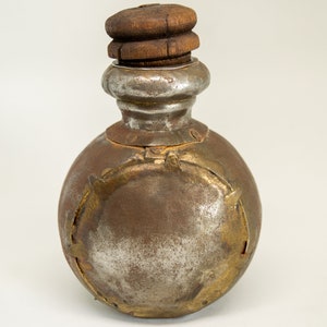 K&ouml;nnte beinhalten: Eine antike Metallflasche mit einem Holzverschluss. Die Flasche ist rund und hat eine silberne Oberfl&auml;che mit goldenen Akzenten. Die Flasche ist leicht angelaufen und zeigt Spuren des Alters.