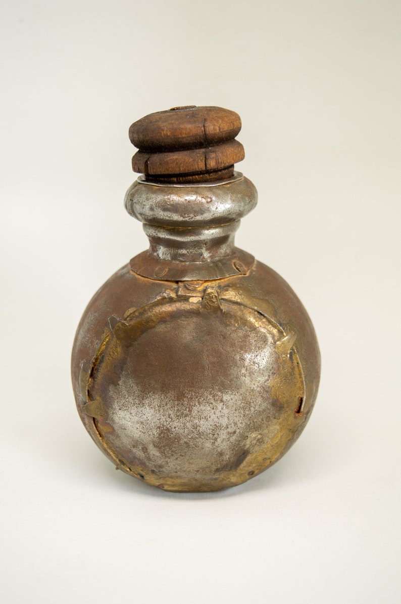 K&ouml;nnte beinhalten: Eine antike Metallflasche mit einem Holzverschluss. Die Flasche ist rund und hat eine silberne Oberfl&auml;che mit goldenen Akzenten. Die Flasche ist leicht angelaufen und zeigt Spuren des Alters.