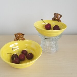 May include: Two yellow ceramic bowls with brown bear figurines on the rims, filled with fresh cherries. One bowl rests on a clear glass pedestal. The bowls are on a light-colored wooden surface.