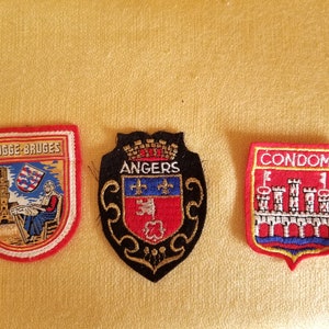 May include: Three embroidered patches with red borders. One reads "BRUGGE-BRUGES" with a scene of a tower and a person. Another says "ANGERS" with a coat of arms. The last says "CONDOM" with a castle.