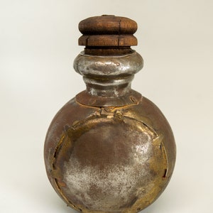 K&ouml;nnte beinhalten: Eine runde, silberne und goldene Metallflasche mit einem Holzdeckel. Die Flasche hat ein dekoratives Design mit goldenen Akzenten.