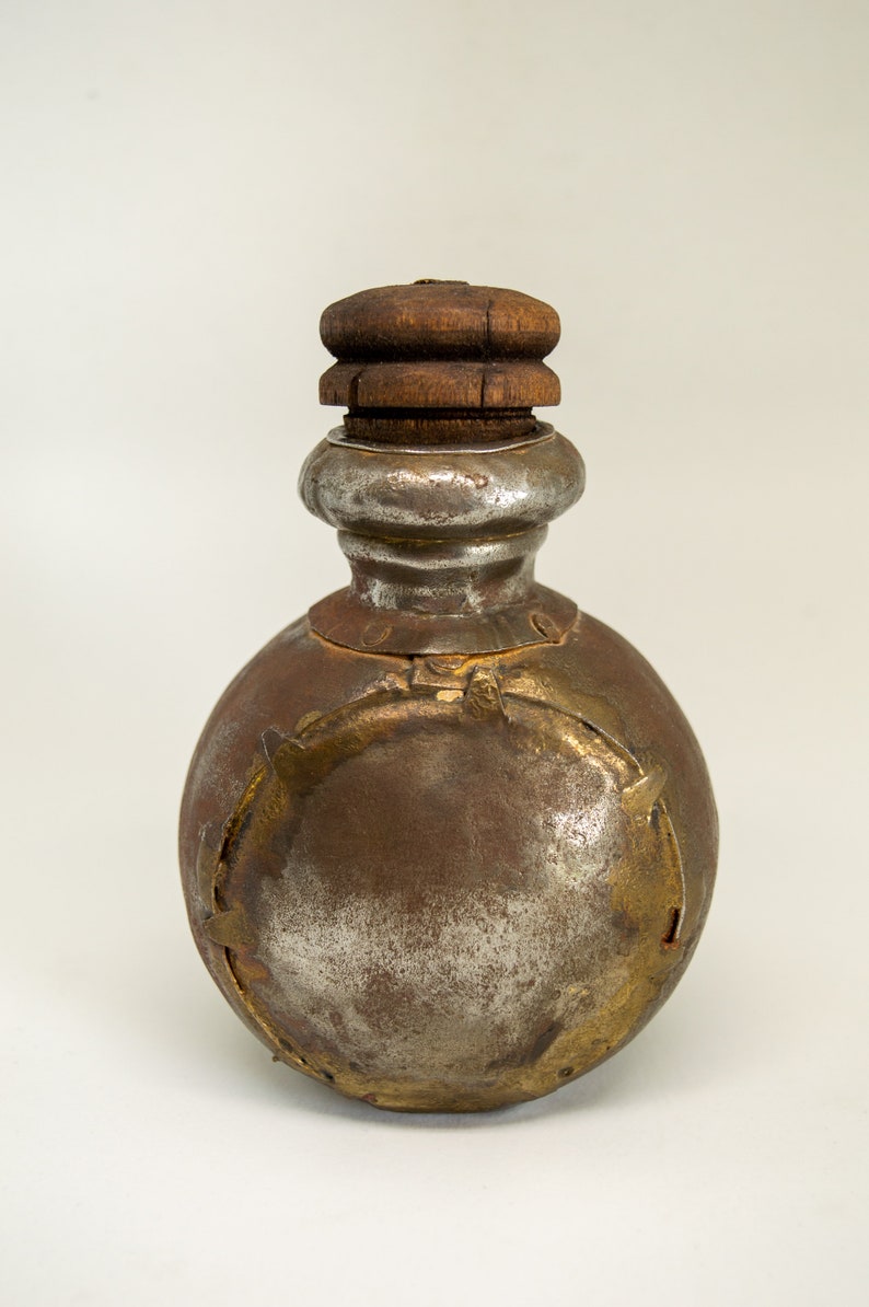 K&ouml;nnte beinhalten: Eine runde, silberne und goldene Metallflasche mit einem Holzdeckel. Die Flasche hat ein dekoratives Design mit goldenen Akzenten.