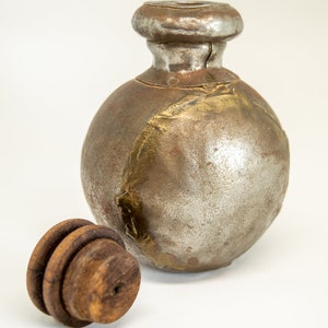 K&ouml;nnte beinhalten: Eine runde, silberne und goldene Metallflasche mit einem Holzstopfen. Die Flasche hat ein abgenutztes, altes Aussehen.
