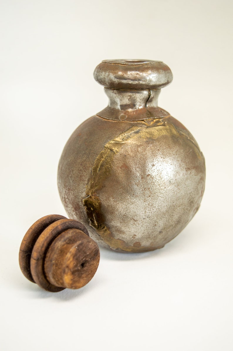 K&ouml;nnte beinhalten: Eine runde, silberne und goldene Metallflasche mit einem Holzstopfen. Die Flasche hat ein abgenutztes, altes Aussehen.