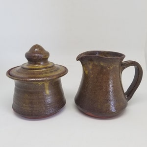 May include: A brown and yellow ceramic creamer and sugar bowl set. The creamer has a rounded body and a handle. The sugar bowl has a lid with a rounded top.