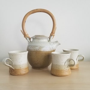 May include: A set of four white and brown speckled ceramic mugs with a matching teapot with a bamboo handle. The teapot and mugs have a speckled glaze with a brown band around the bottom.