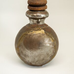 K&ouml;nnte beinhalten: Eine kleine, runde Metallflasche mit einem Holzstopfen. Die Flasche ist rostig und hat eine silberne Oberfl&auml;che. Die Flasche ist mit goldenen Akzenten verziert.