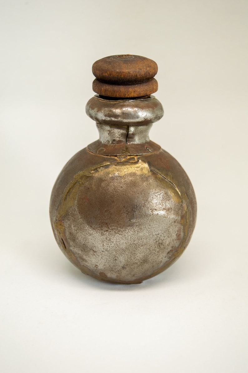 K&ouml;nnte beinhalten: Eine kleine, runde Metallflasche mit einem Holzstopfen. Die Flasche ist rostig und hat eine silberne Oberfl&auml;che. Die Flasche ist mit goldenen Akzenten verziert.