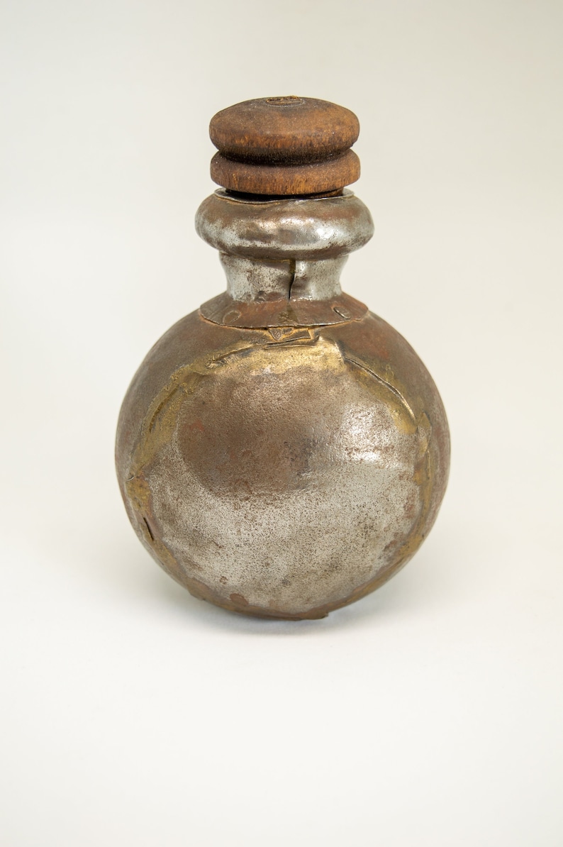 K&ouml;nnte beinhalten: Eine runde, silberne und braune Metallflasche mit einem Holzstopfen. Die Flasche hat ein rustikales, altes Aussehen.
