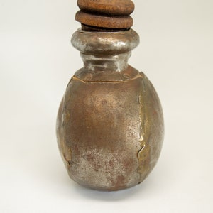 K&ouml;nnte beinhalten: Eine kleine, runde Metallflasche mit einem Holzstopfen. Die Flasche ist rostbraun mit silbernen Akzenten.