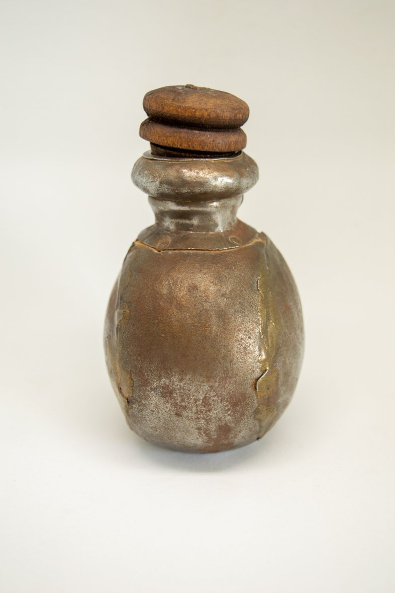 K&ouml;nnte beinhalten: Eine kleine, runde Metallflasche mit einem Holzstopfen. Die Flasche ist rostbraun mit silbernen Akzenten.