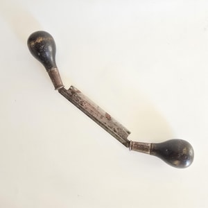 Könnte beinhalten: Ein Zugmesser mit einer langen, geraden Metallklinge mit Gebrauchsspuren und Oxidation, die an beiden Enden durch silberfarbene Metallzwingen mit zwei dunklen, abgerundeten Holzgriffen verbunden ist. Das Werkzeug liegt diagonal.
