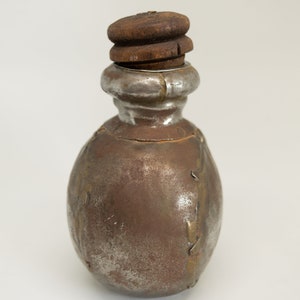 K&ouml;nnte beinhalten: Eine kleine, runde Metallflasche mit einem Holzstopfen. Die Flasche ist silbrig mit braunen Akzenten und wirkt alt oder antik.