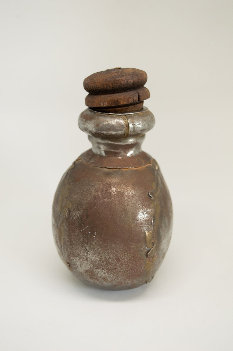 K&ouml;nnte beinhalten: Eine kleine, runde Metallflasche mit einem Holzstopfen. Die Flasche ist silbrig mit braunen Akzenten und wirkt alt oder antik.