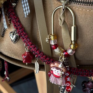 May include: A brass carabiner keychain adorned with red, pink, and clear beads, a small cowboy character charm, a pink tassel, and letter beads spelling HOWDY. A braided red and black cord features silver heart, star, and feather charms.