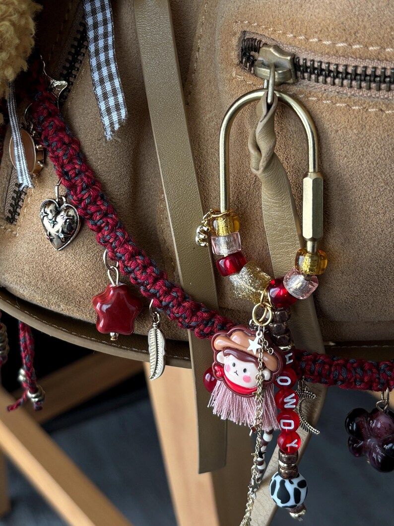 May include: A brass carabiner keychain adorned with red, pink, and clear beads, a small cowboy character charm, a pink tassel, and letter beads spelling HOWDY. A braided red and black cord features silver heart, star, and feather charms.