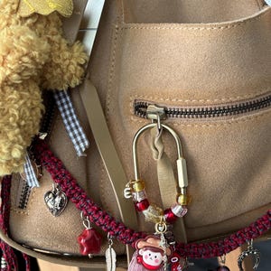 May include: A tan suede bag adorned with a gold-toned carabiner keychain featuring red, yellow, and clear beads, a small character charm, and a pink tassel. The bag also features a braided red and black strap with various metal charms including a heart, star, feather, and crescent moon.