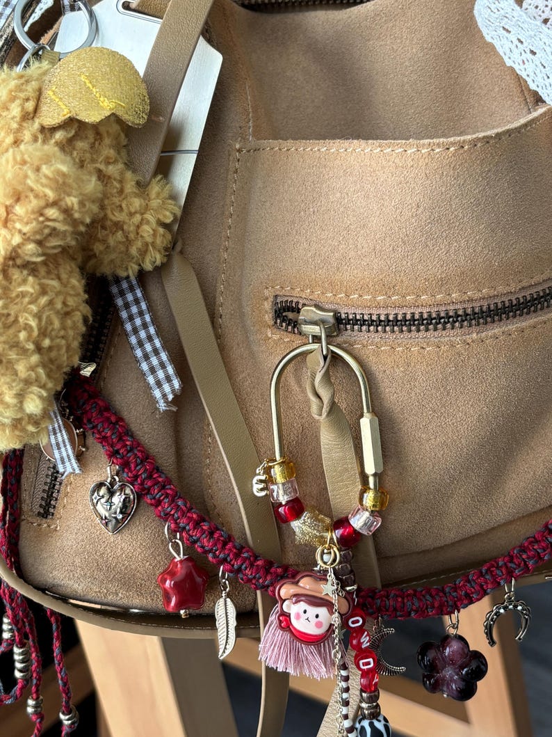May include: A tan suede bag adorned with a gold-toned carabiner keychain featuring red, yellow, and clear beads, a small character charm, and a pink tassel. The bag also features a braided red and black strap with various metal charms including a heart, star, feather, and crescent moon.