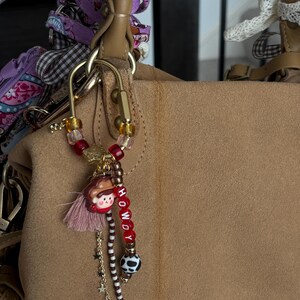 May include: A gold-toned metal carabiner keychain featuring a variety of beads, including red, clear, and brown patterned spheres. It includes a small charm of a person wearing a brown hat, a pink tassel, a gold chain with star charms, and red letter beads spelling HOWDY.