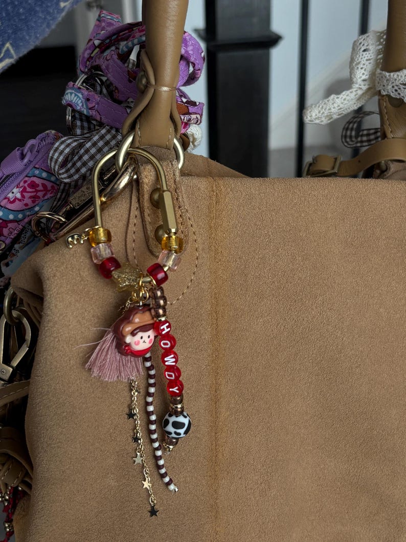 May include: A gold-toned metal carabiner keychain featuring a variety of beads, including red, clear, and brown patterned spheres. It includes a small charm of a person wearing a brown hat, a pink tassel, a gold chain with star charms, and red letter beads spelling HOWDY.