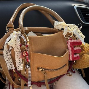 May include: A tan suede handbag featuring a front zippered pocket, two top handles, and decorative accents including white lace ribbons, a beaded letter E charm, and various small dangling charms with red and brown details attached to the hardware.