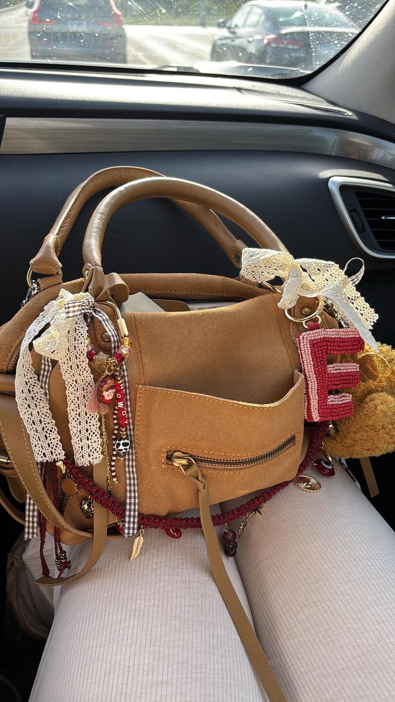 May include: A tan suede handbag featuring a front zippered pocket, two top handles, and decorative accents including white lace ribbons, a beaded letter E charm, and various small dangling charms with red and brown details attached to the hardware.